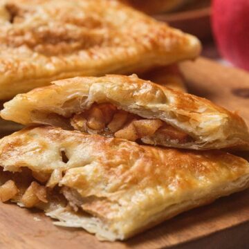 Flaky pastry turnovers filled with diced apples on a wooden board, with whole apples in the background.