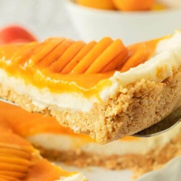 A close-up of a slice of mango cheesecake with a crumbly crust held up on a spatula.
