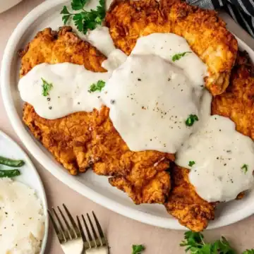 Fried chicken cutlets topped with white gravy on a platter, served with green beans and mashed potatoes.