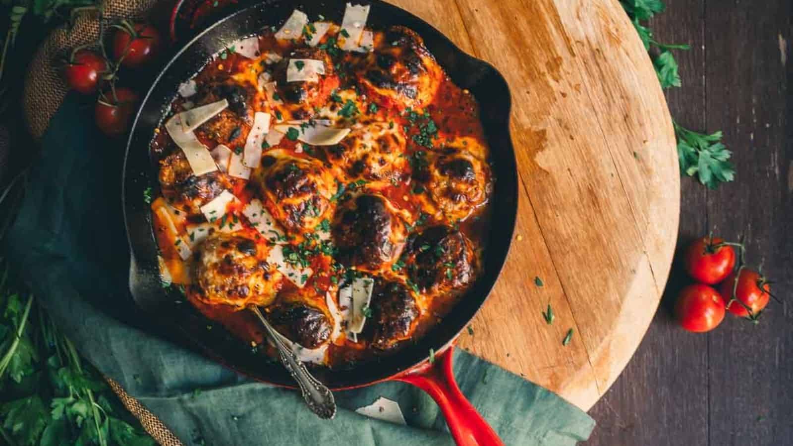 Meatballs in tomato sauce topped with parmesan in a skillet on a wooden board, garnished with herbs.