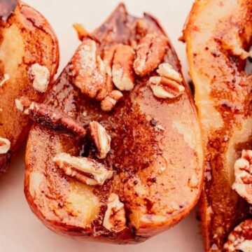 Three baked pear halves topped with chopped pecans and cinnamon on a white surface.