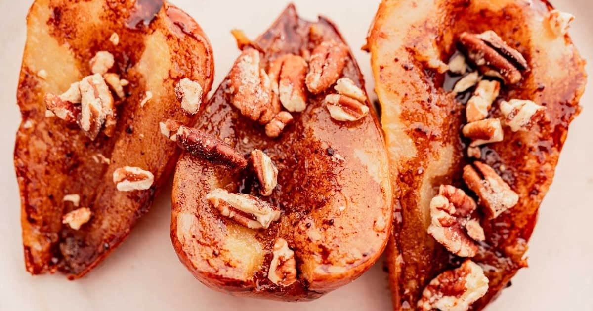 Three baked pear halves topped with chopped pecans and cinnamon on a white surface.