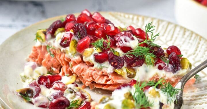 Salmon fillet topped with yogurt sauce, pomegranate seeds, pistachios, and fresh dill on a plate.