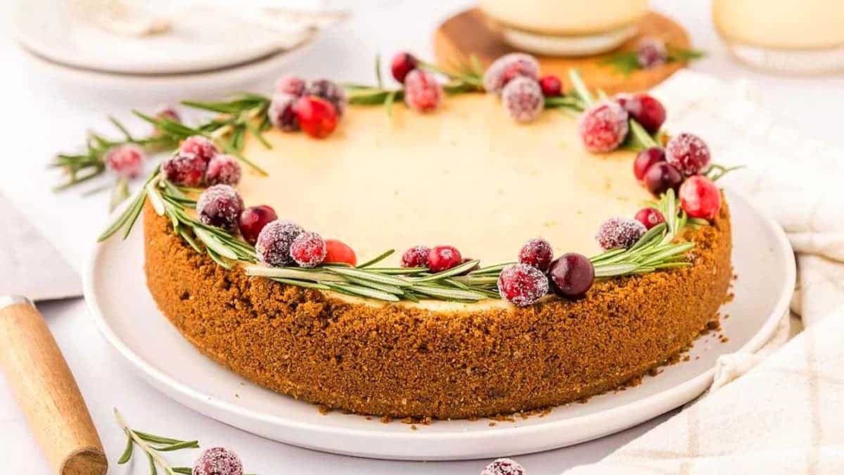 Cheesecake topped with sugared cranberries and rosemary on a white plate, with a golden-brown crumb crust.