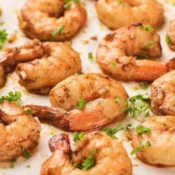 Seasoned, cooked shrimp garnished with parsley and lemon wedges on a white surface.