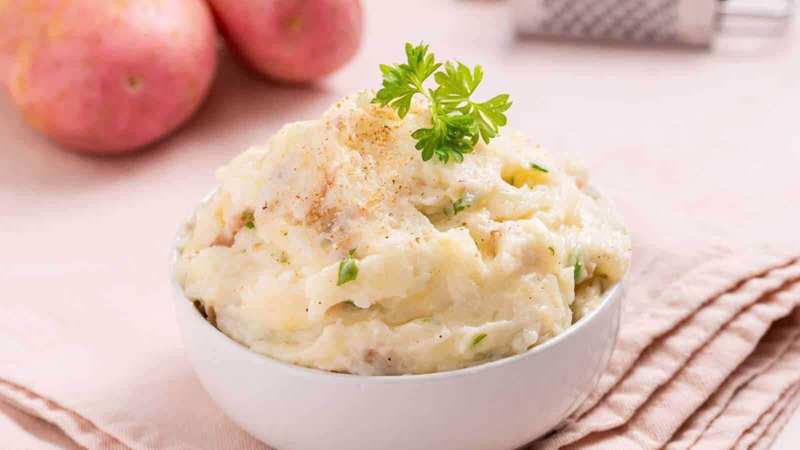 A bowl of mashed potatoes garnished with parsley, set on a pink cloth napkin, with whole potatoes and a grater in the background.
