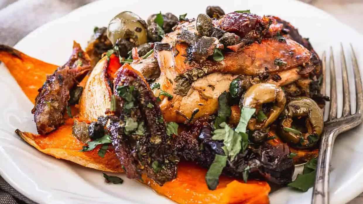 Chicken with olives, capers, and herbs on roasted sweet potato wedges makes a flavorful addition to your thanksgiving mains, served elegantly on a white plate with a fork.