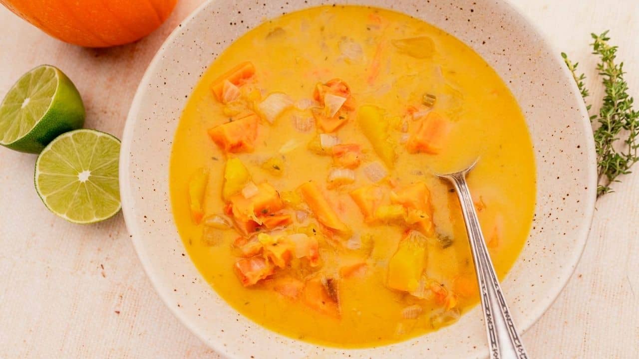 Bowl of orange vegetable soup with a spoon, next to lime halves and fresh herbs on a light surface.