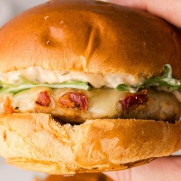 A hand holding a sandwich with lettuce, sauce, melted cheese, and bacon on a brioche bun.