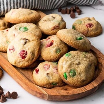 A wooden tray of cookies with red and green candies, chocolate chips nearby, on a marble surface.