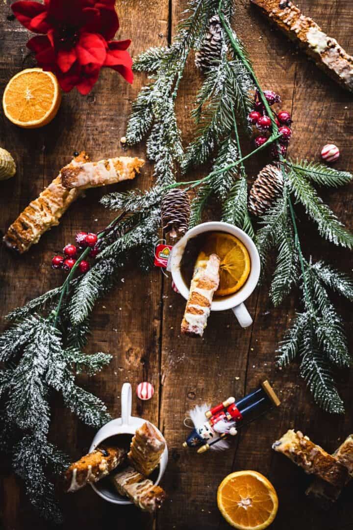 Snowy pine branches, Pistachio Gluten-Free Biscotti, and mugs with orange slices on a rustic wooden table with festive decorations.