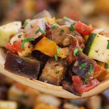 A wooden spoon holds colorful diced vegetables like zucchini, eggplant, tomato, and peppers garnished with herbs.