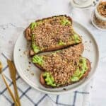 Two slices of Avocado Toast With Dukkah are topped with seasoning on a white plate, next to gold utensils on a plaid cloth.