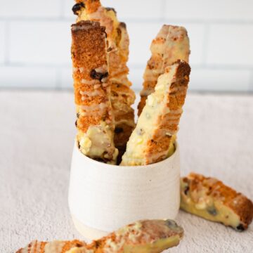 Four Pistachio Gluten-Free Biscotti dipped in white chocolate stand upright in a white cup, with two more biscotti on the table.