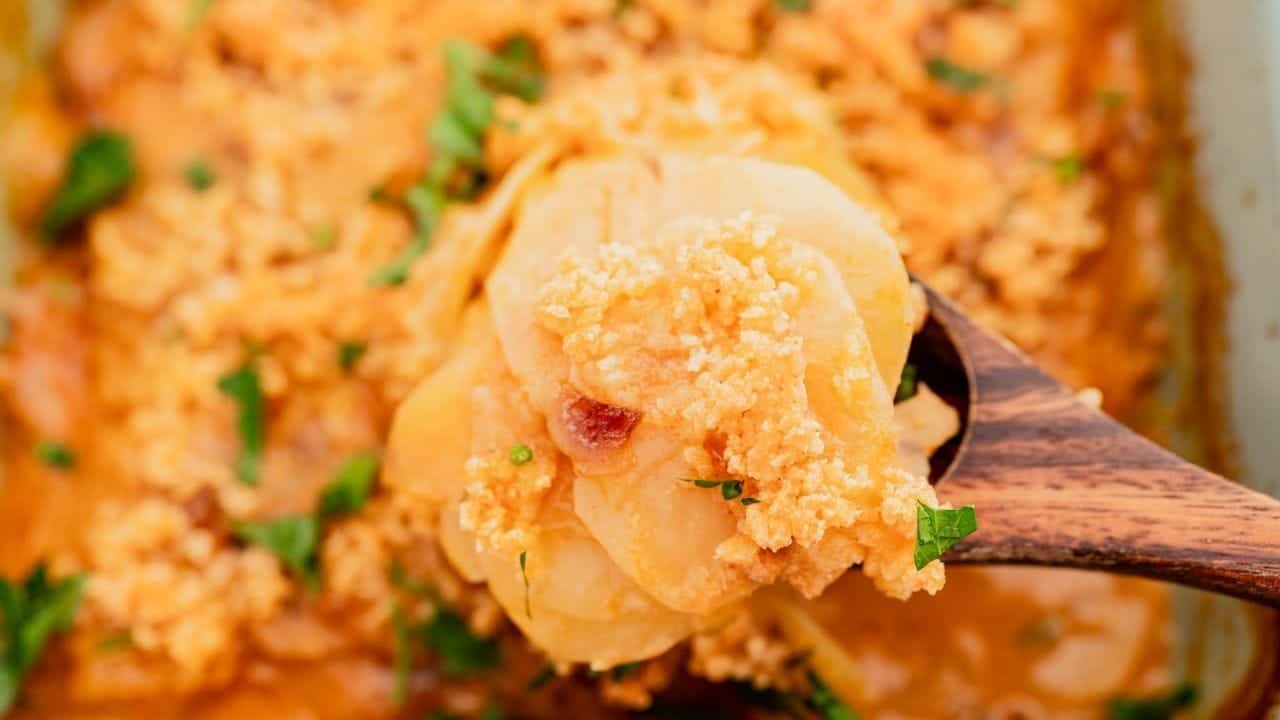 A wooden spoon holds creamy, cheesy sliced potatoes topped with herbs and sauce.
