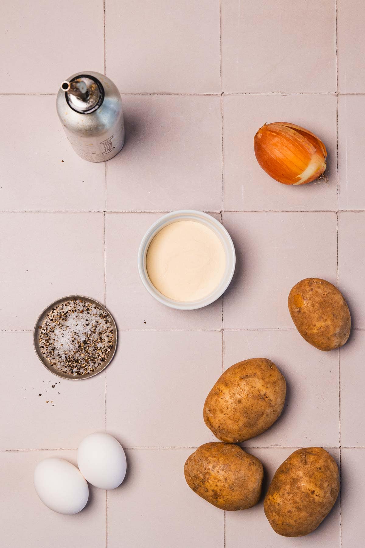Potatoes, eggs, onion, cream, black pepper, and a metal canister arranged on a tiled surface.