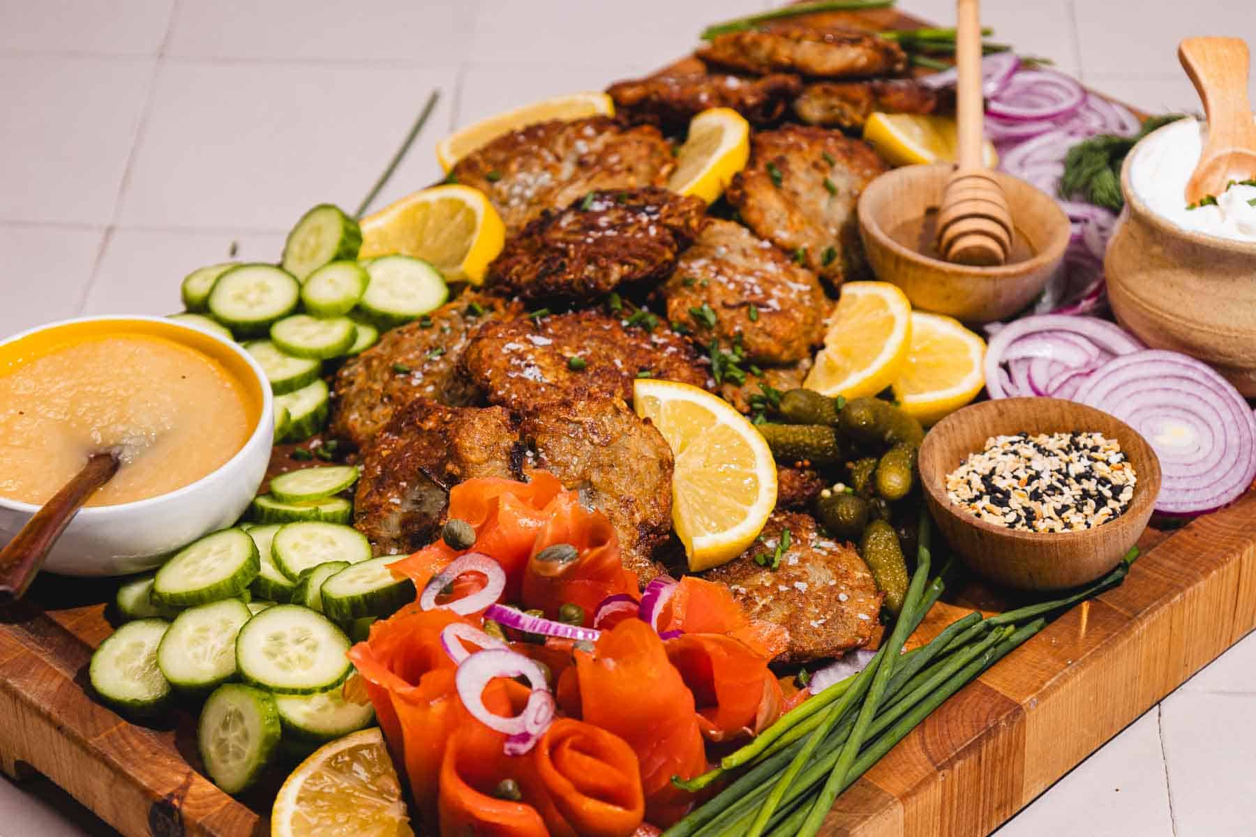 A wooden platter with latkes, lemon slices, cucumber, smoked salmon, dip, and garnishes.