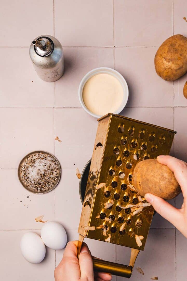 Hands grating a potato over a box grater, with eggs, cream, pepper, and a whipped cream dispenser nearby.