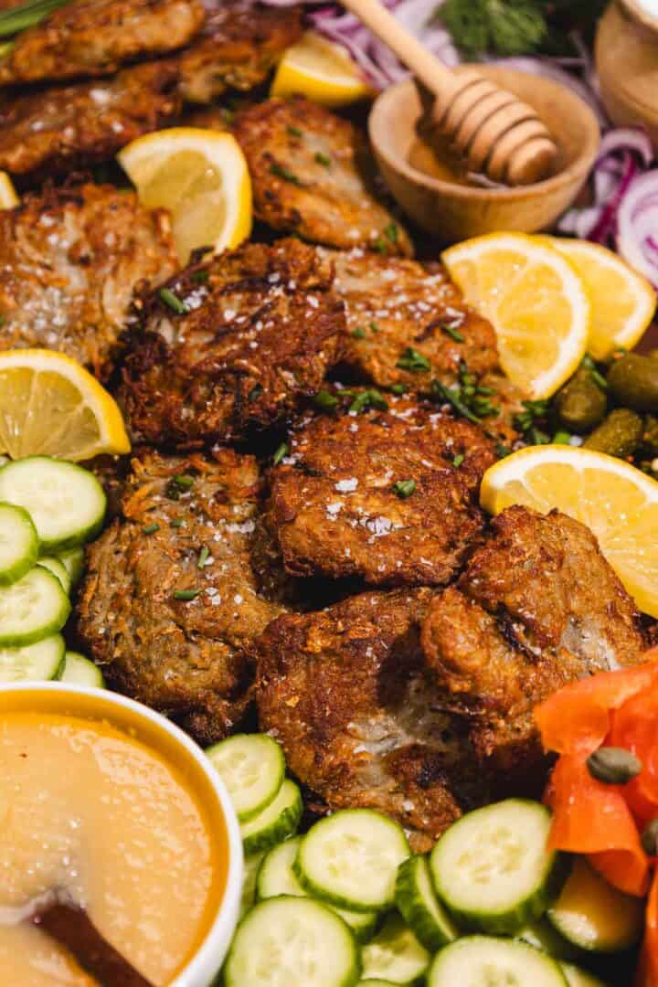 Crispy latkes surrounded by cucumber slices, lemon wedges, applesauce, and garnishes on a platter.