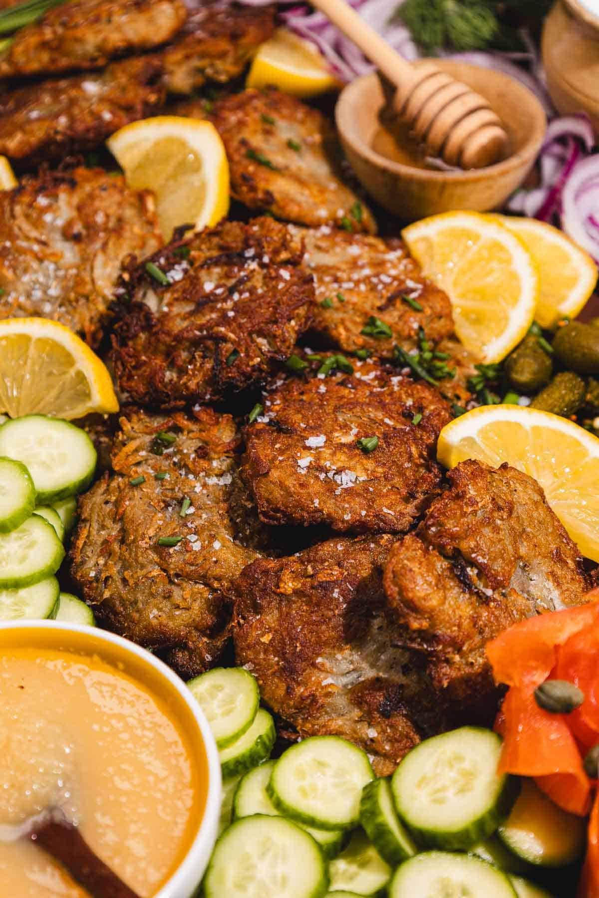Crispy latkes surrounded by cucumber slices, lemon wedges, applesauce, and garnishes on a platter.