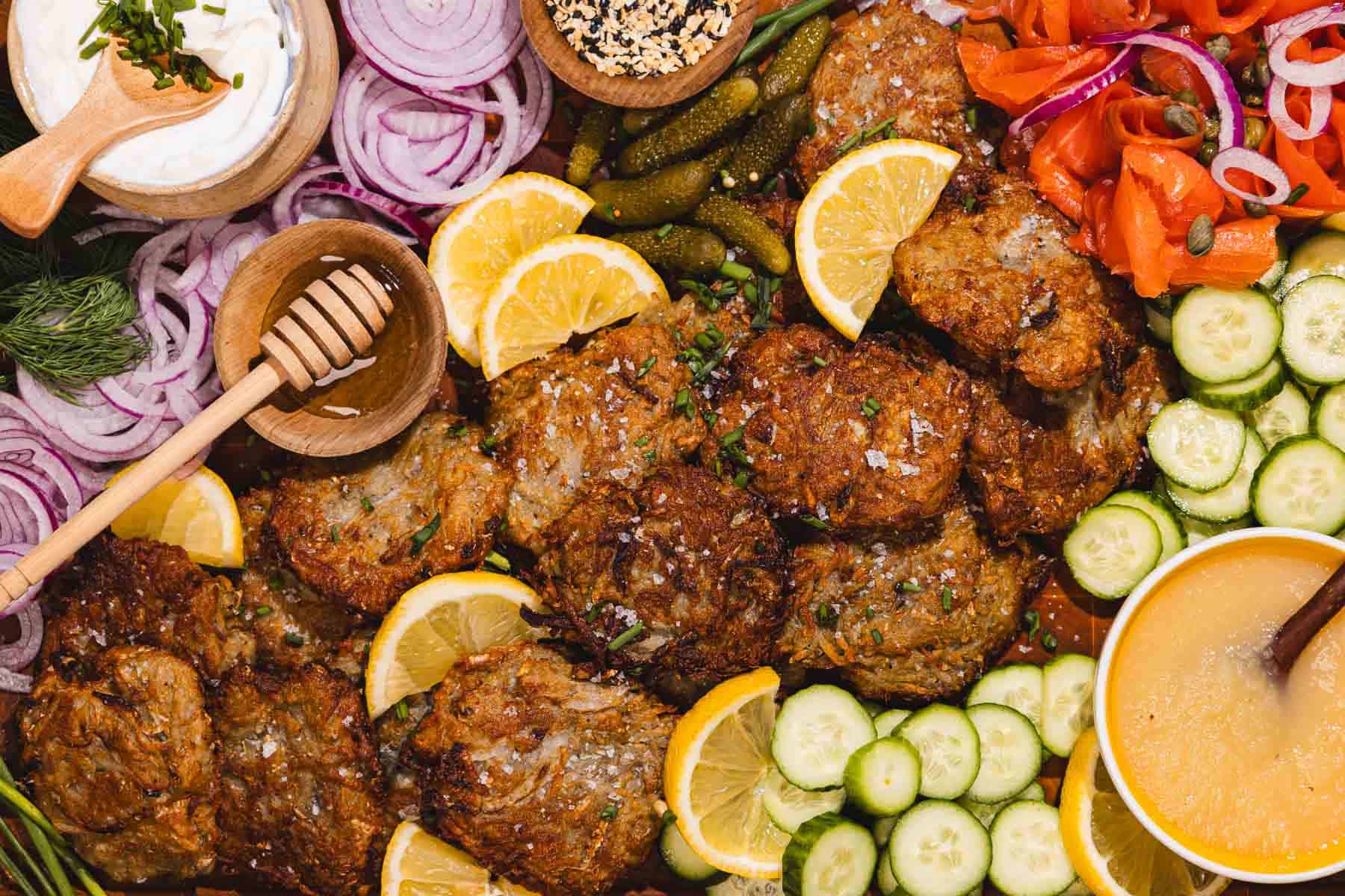 Crispy latkes with lemon slices, cucumber, smoked salmon, onions, honey, and dipping sauces on a platter.