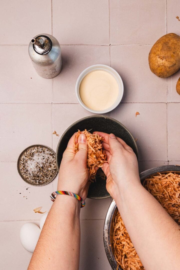 Hands squeezing shredded potatoes over a bowl, surrounded by eggs, spices, cream, and whole potatoes.