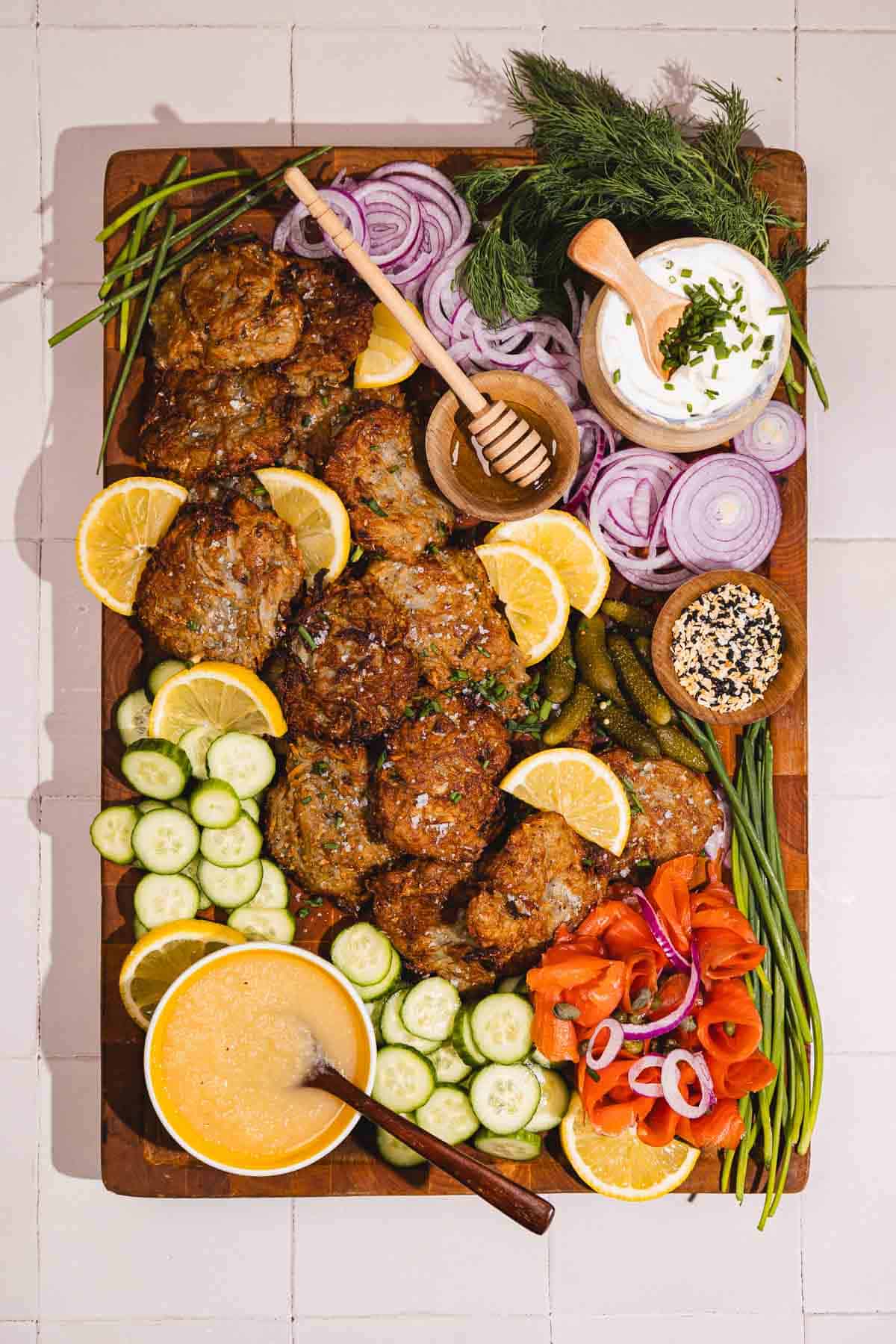 A wooden board with fritters, sliced cucumbers, lemon, red onions, sauce, herbs, and pickled vegetables.