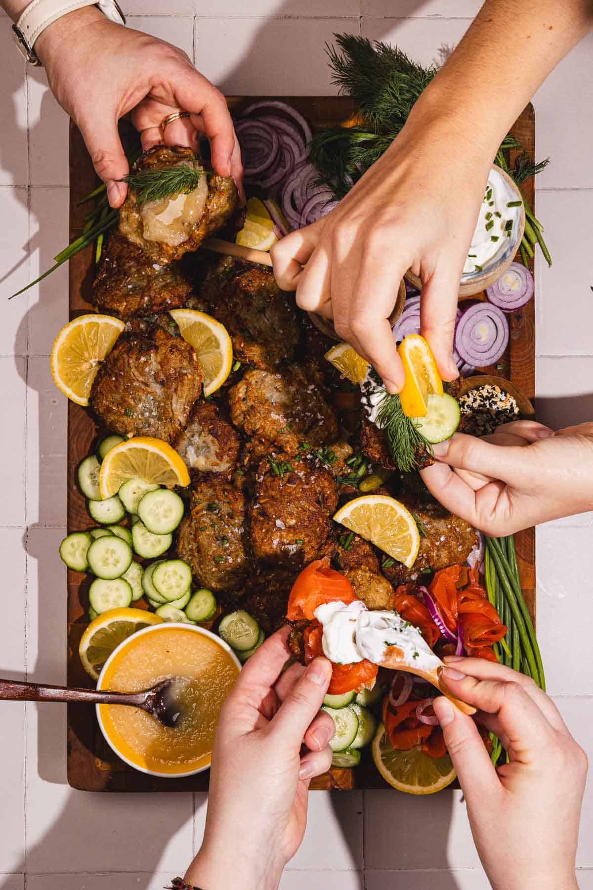 Four hands reaching for falafel, veggies, and dips on a platter garnished with lemon slices and herbs.