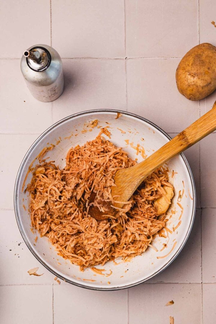 Bowl of grated potatoes mixed with spices, a wooden spoon, a raw potato, and oil spray on a tiled surface.
