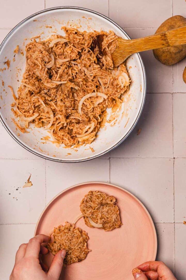 A hand shapes potato-onion mixture from a bowl onto a pink plate on a tiled surface.