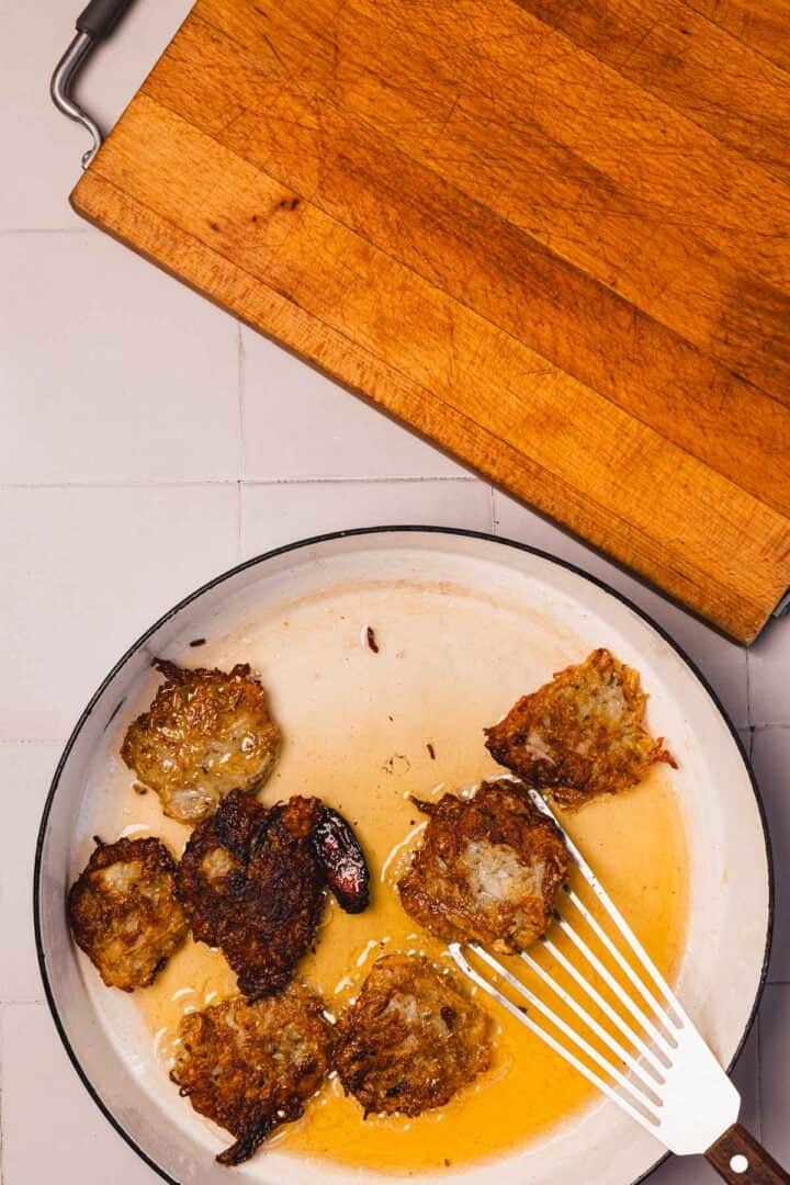 Seven fried latkes in a pan with oil, spatula resting inside, wooden cutting board nearby.