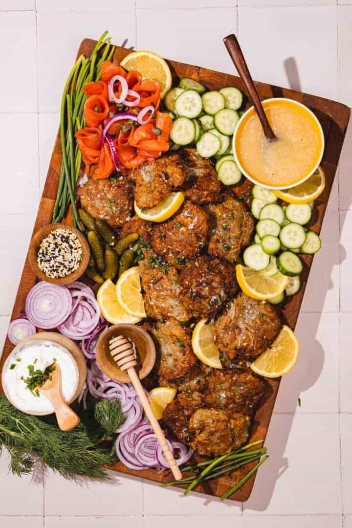A wooden platter with latkes, lemon wedges, sliced cucumbers, red onions, lox, dip, pickles, and garnishes.