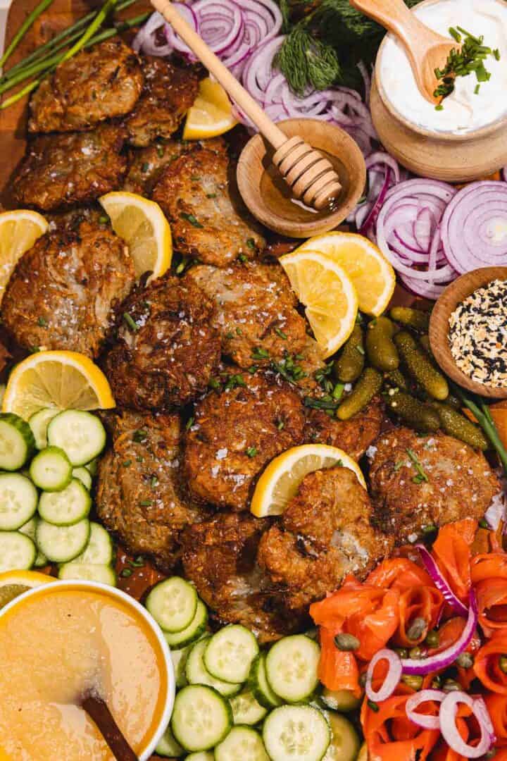 A platter of crispy latkes with lemon slices, cucumber, pickles, onions, sour cream, honey, and vegetables.
