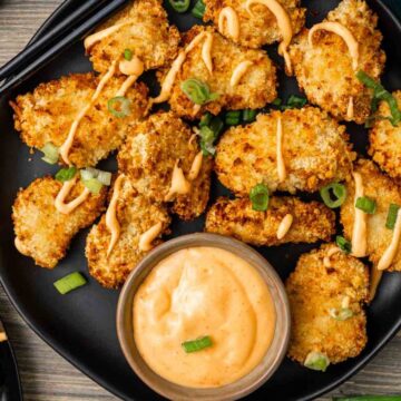 A black plate of fried breaded mushrooms drizzled with sauce, garnished with green onions, and a dip.