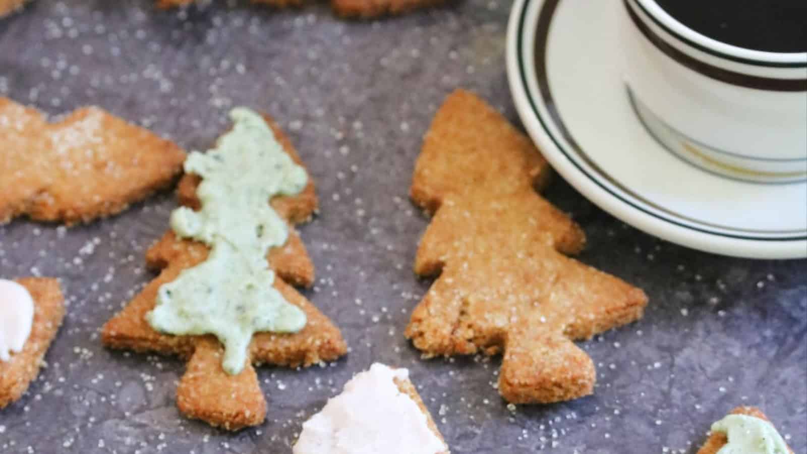 Christmas tree-shaped cookies with icing next to a cup of coffee on a dark surface.