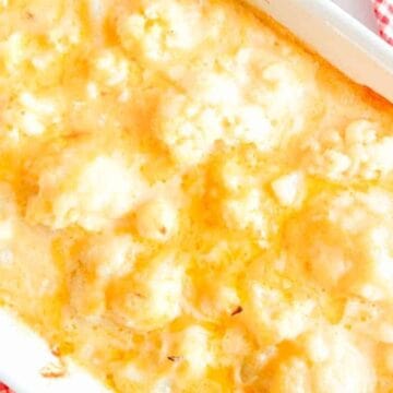 Baked cauliflower cheese in a white casserole dish on a red and white checkered cloth, with a spoon nearby.