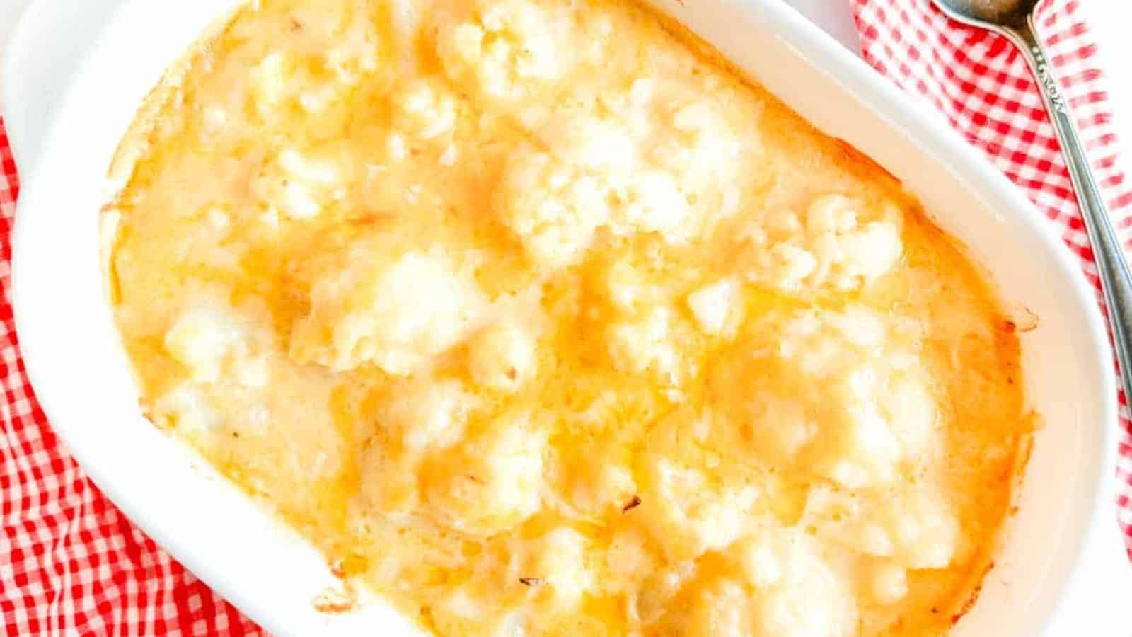 Baked cauliflower cheese in a white casserole dish on a red and white checkered cloth, with a spoon nearby.