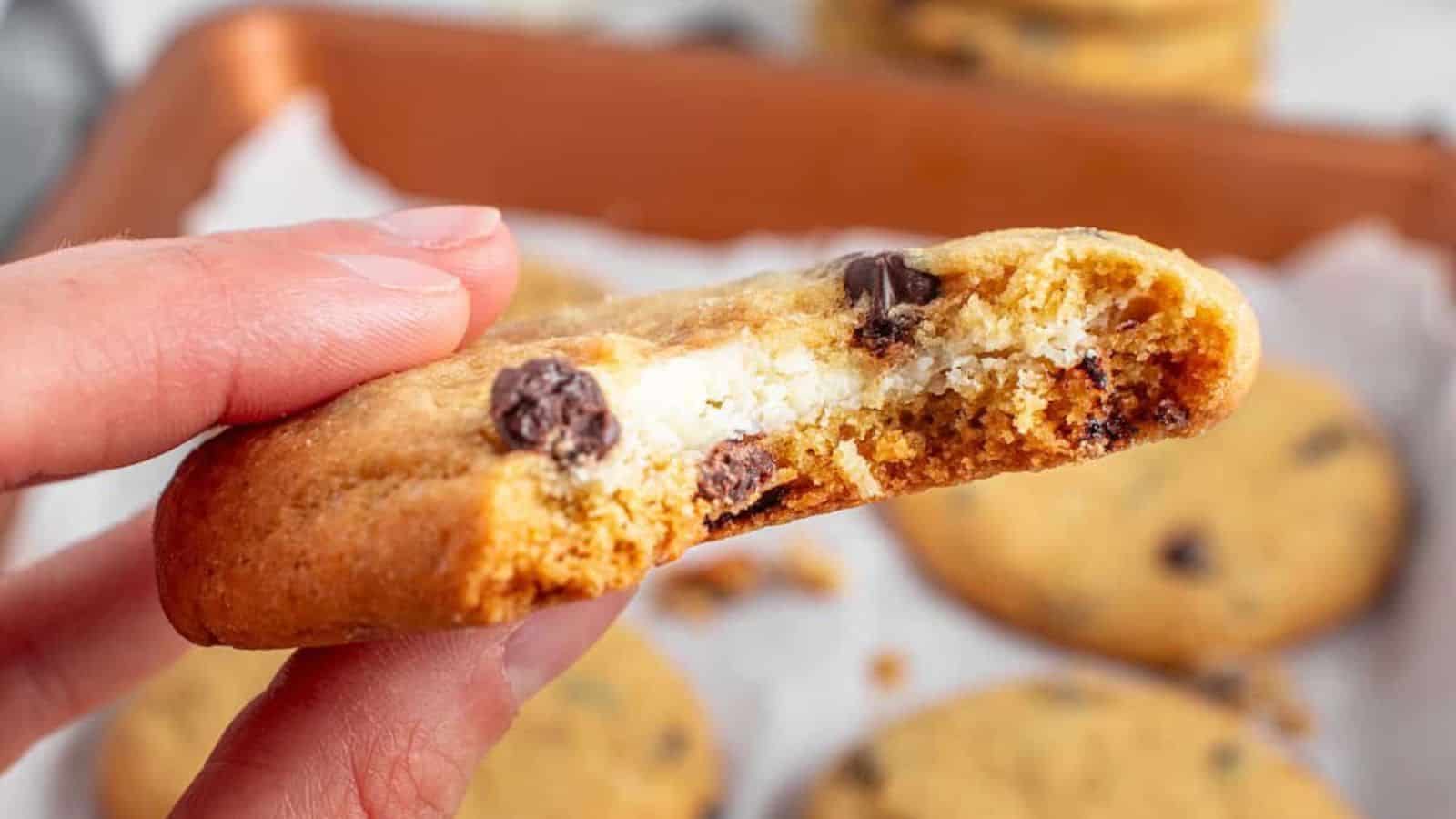 A hand holding a half-eaten chocolate chip cookie with a cheesecake filling. Other cookies are visible on a baking tray in the background.