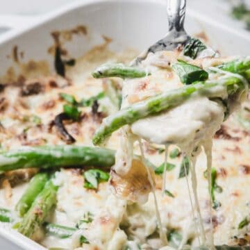 A cheesy green bean casserole being served from a white baking dish with a spoon.
