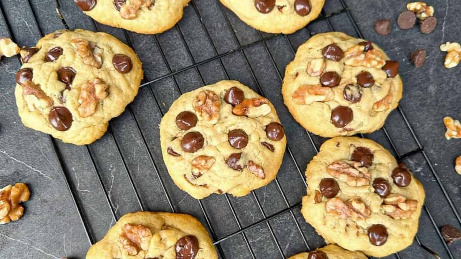 Chocolate chip and walnut cookies cooling on a black wire rack over a dark surface.
