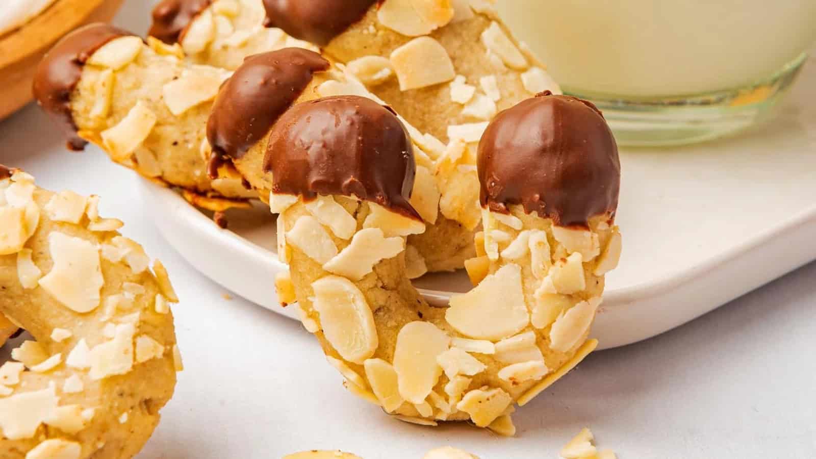 Half-moon almond cookies dipped in chocolate are arranged on a white plate next to a glass of milk.