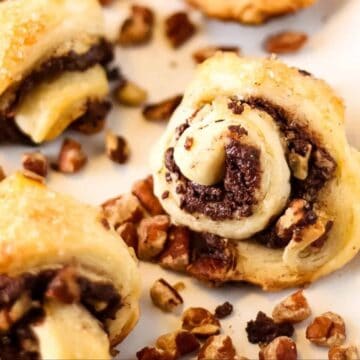 Close-up of chocolate and nut rugelach pastries on parchment paper, surrounded by chopped nuts.