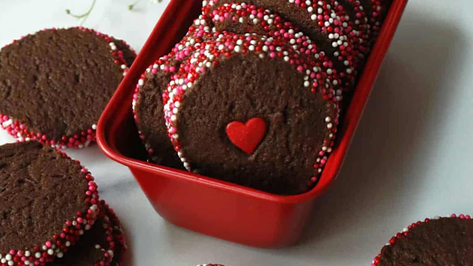 Chocolate cookies with red, pink, and white sprinkles in a red container, one with a heart decoration.