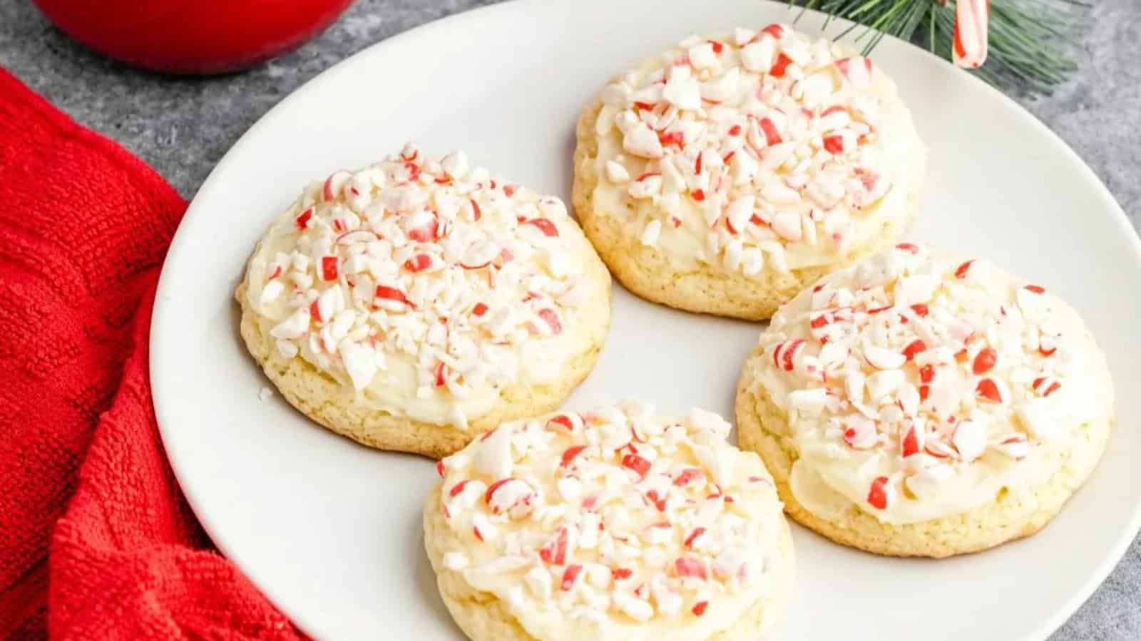 Four frosted sugar cookies topped with crushed peppermint on a white plate, next to a red cloth and greenery.