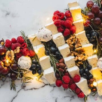 Cheese, grapes, berries, nuts, and herbs arranged in the shape of a Christmas tree, with string lights.