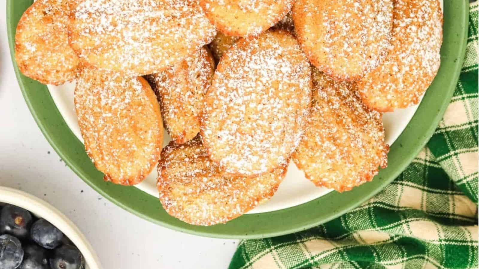 A plate of golden madeleines dusted with powdered sugar, next to a green checkered cloth and blueberries.