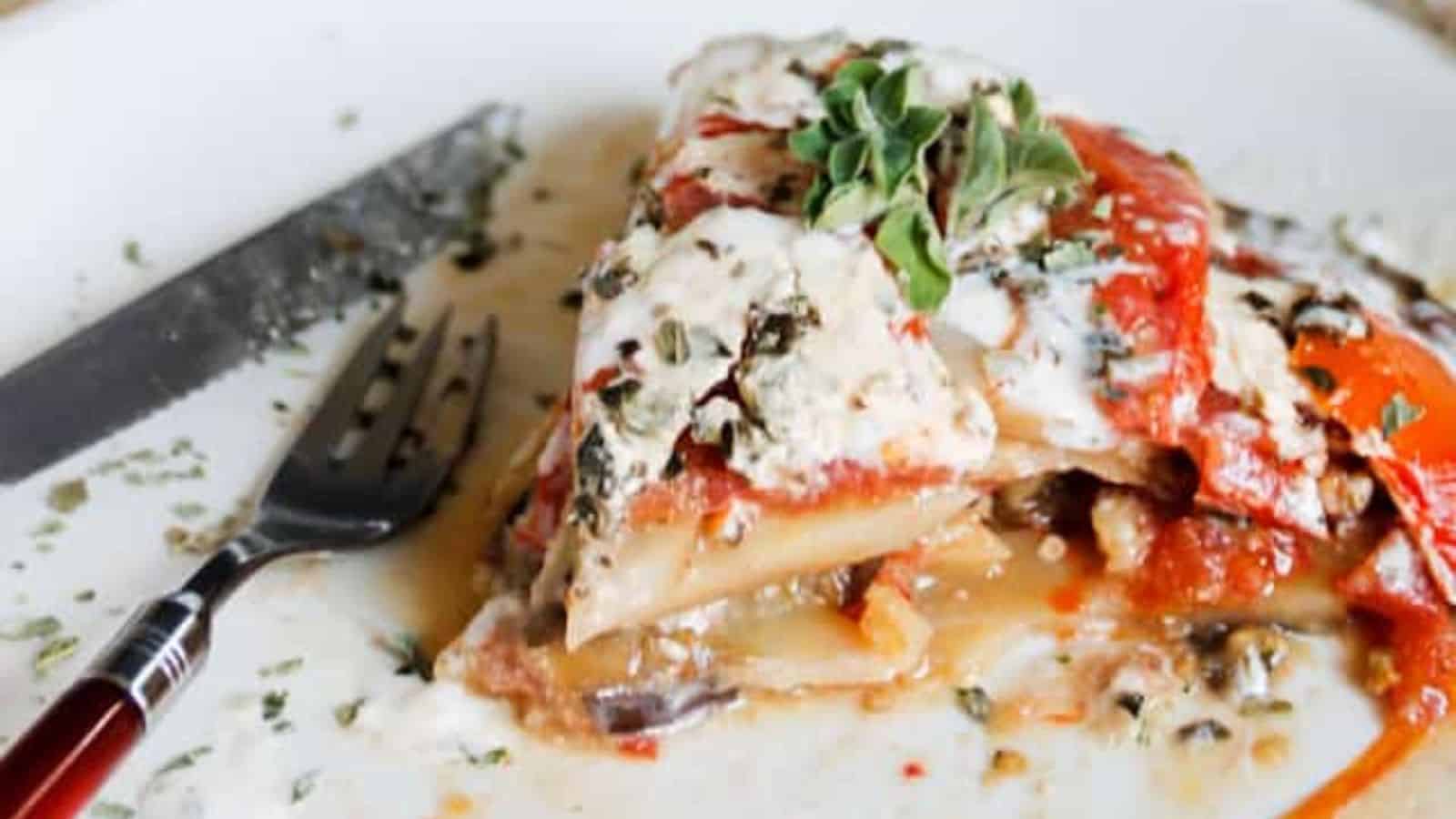A slice of vegetable lasagna topped with herbs and creamy sauce on a white plate with a fork and knife.