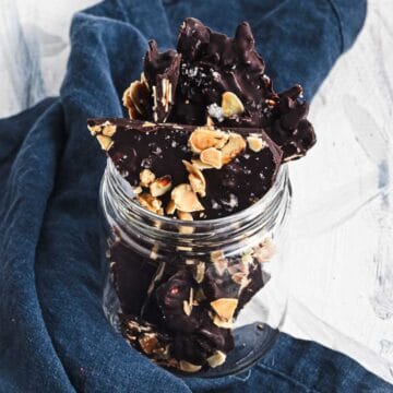 Dark chocolate bark with nuts in a glass jar, placed on a dark blue cloth against a light background.