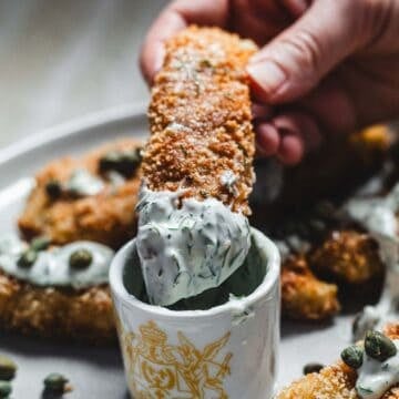 A hand dips a breaded, fried zucchini stick into a cup of creamy herb sauce.