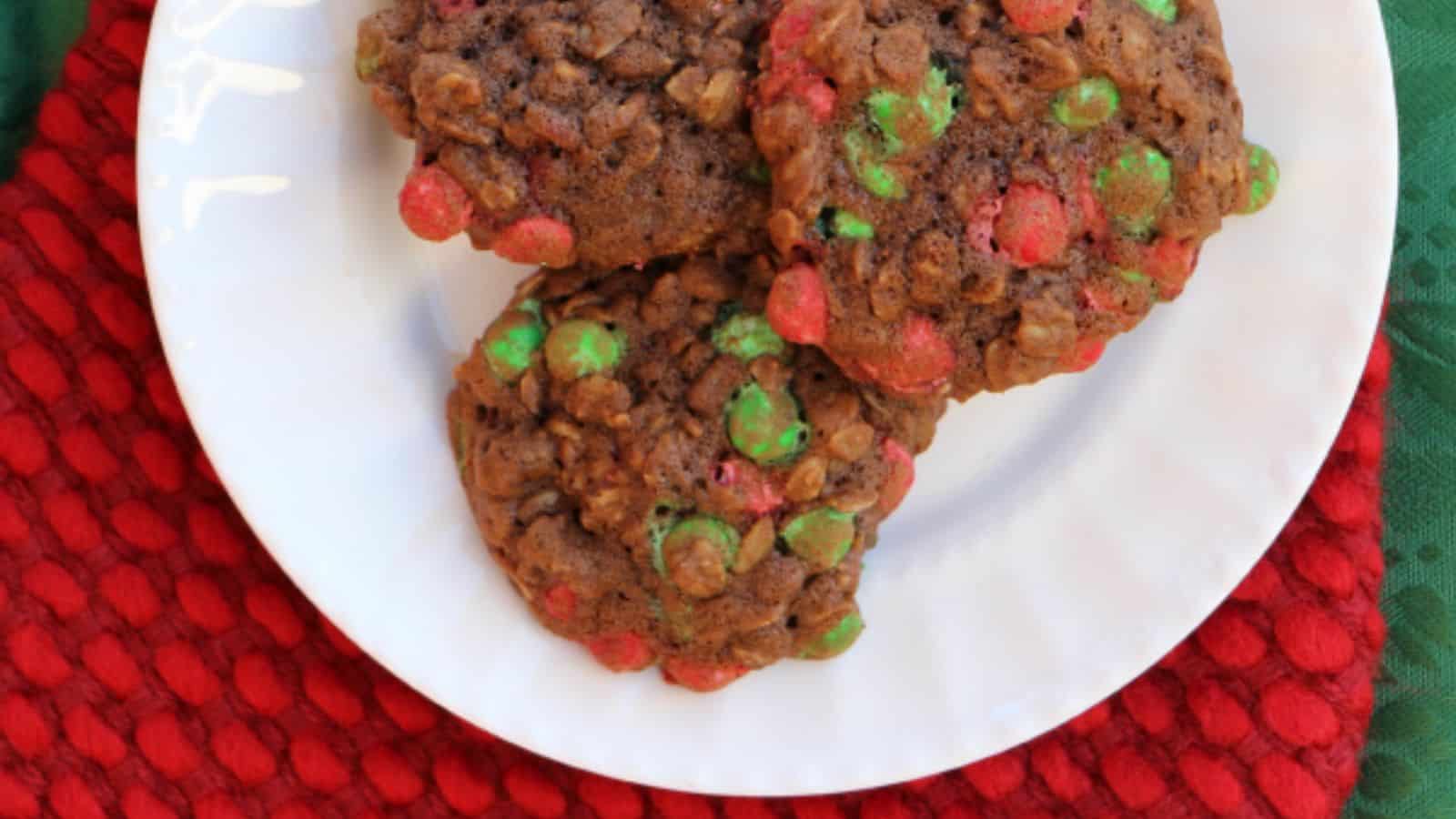Three chocolate cookies with red and green candy pieces on a white plate, atop a red textured cloth.